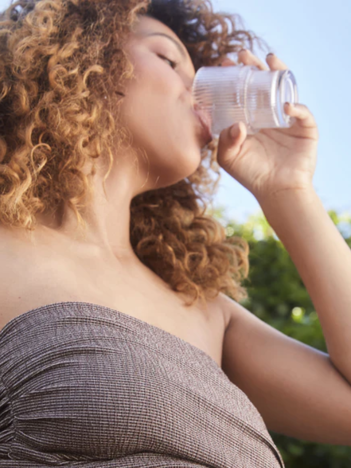 A person with curly hair drinks water from a clear glass outdoors, wearing a strapless top, with foliage visible in the background.