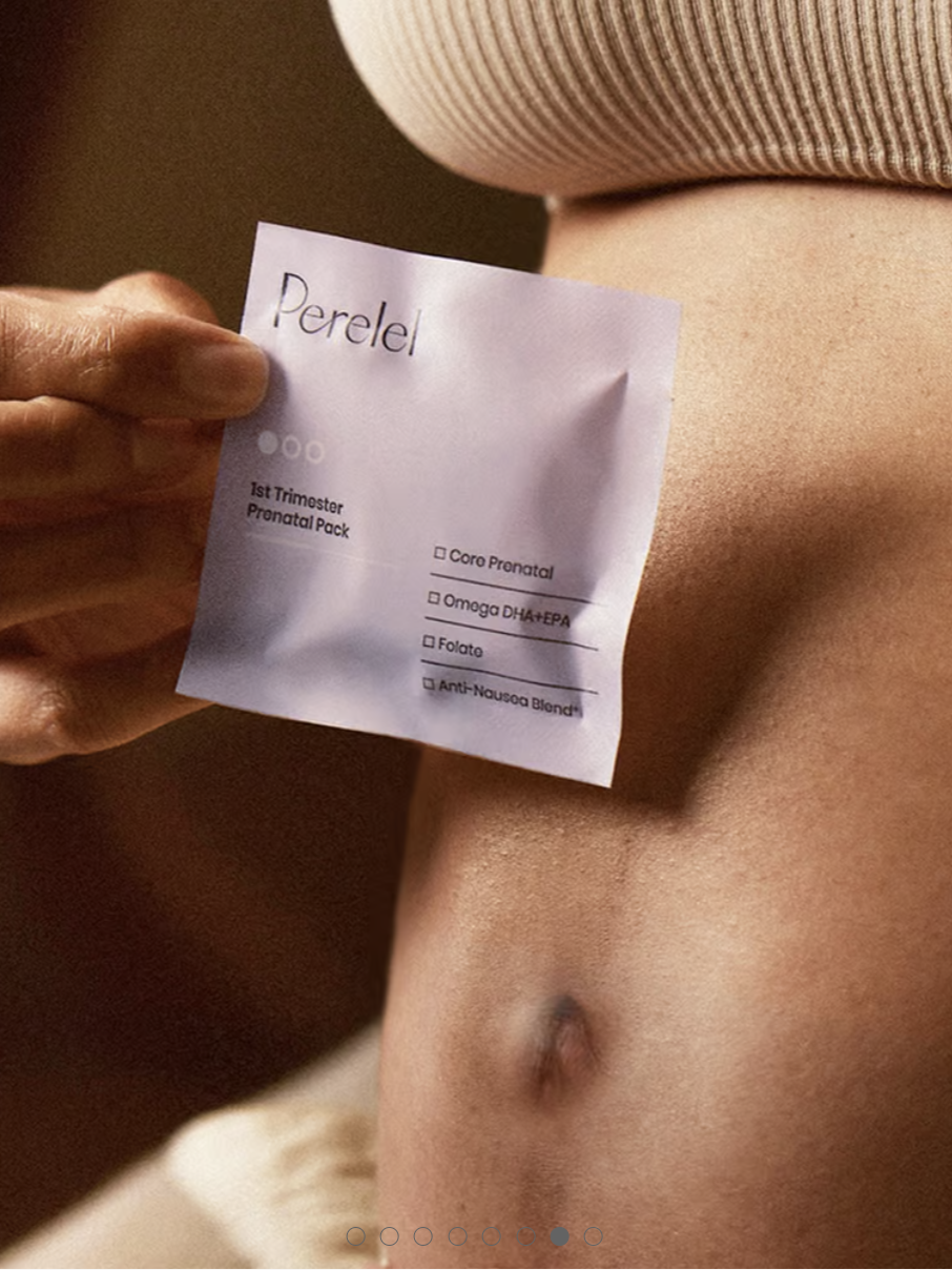 Hand holding a packet labeled "Perelel" against a pregnant belly. The packet lists vitamins for the 1st trimester: Core Prenatal, Omega DHA/EPA, Folate, Anti-Nausea Blend.