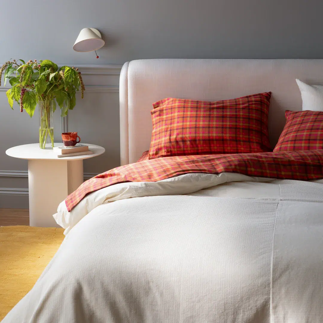 Bedroom with a large bed featuring red plaid pillows and a white comforter, next to a white round side table with a green plant, glass cup, and wall-mounted light above.