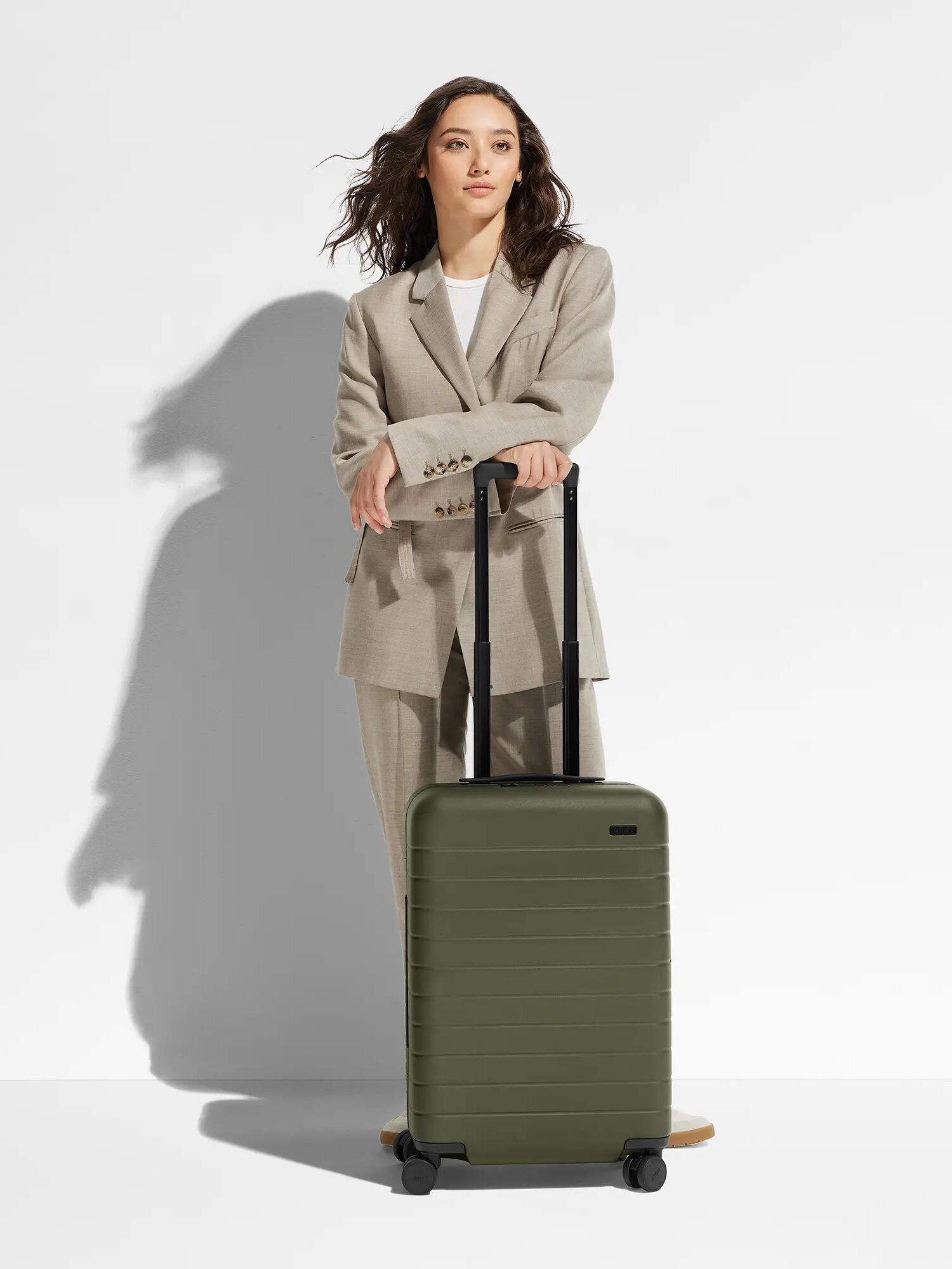 A person in a light gray suit stands against a white background next to an olive green rolling suitcase. Their hand is resting on the suitcase handle.