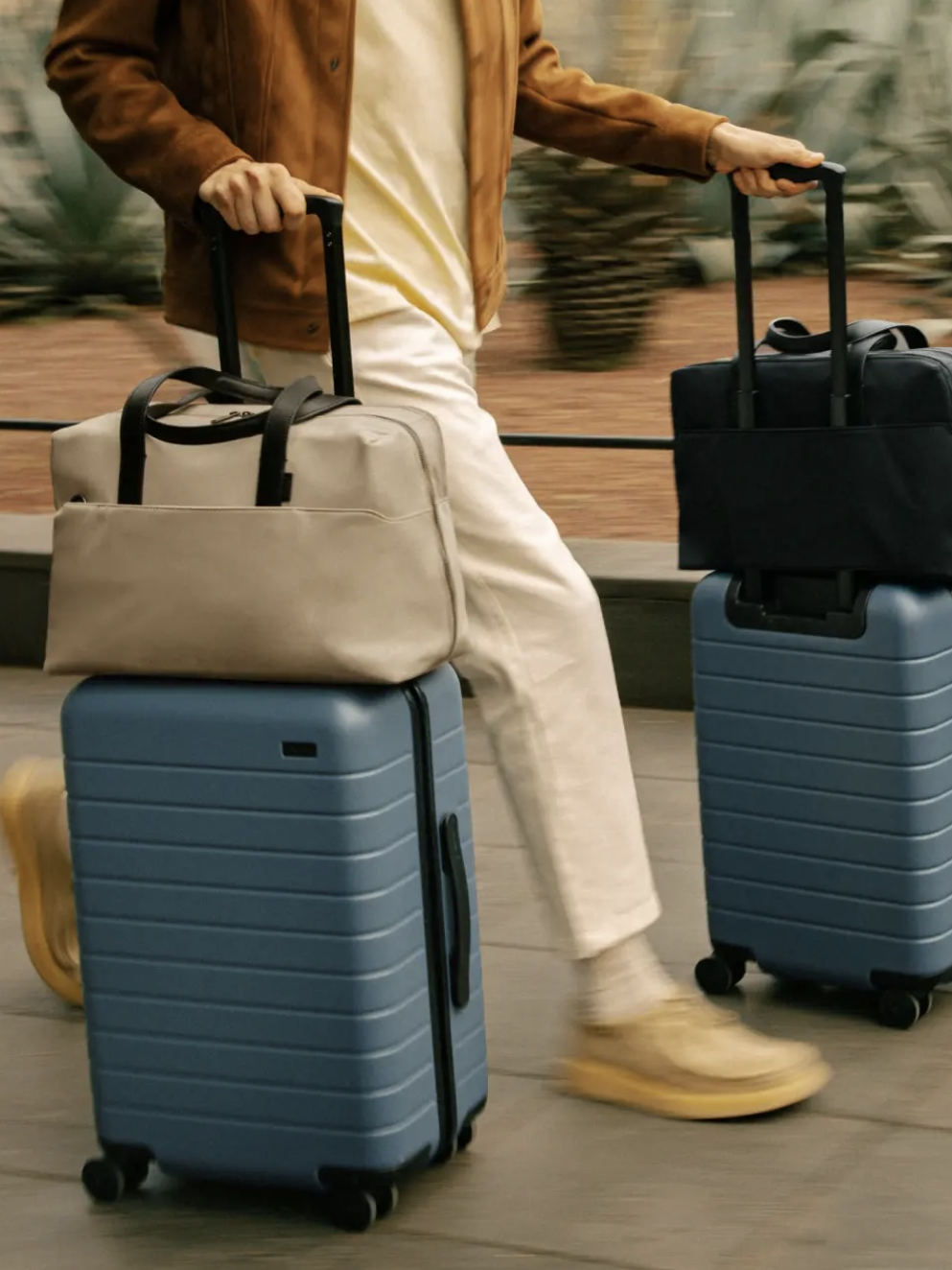 Person walking with two blue rolling suitcases, each topped with a bag, on a paved path near plants.