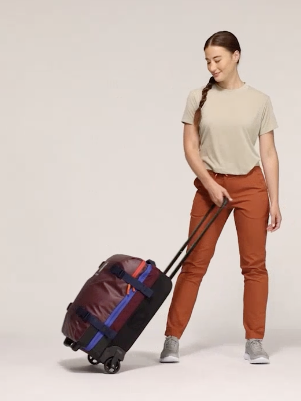 Person with a braided ponytail pulls a wheeled red and black suitcase while wearing a beige shirt, rust-colored pants, and gray sneakers.