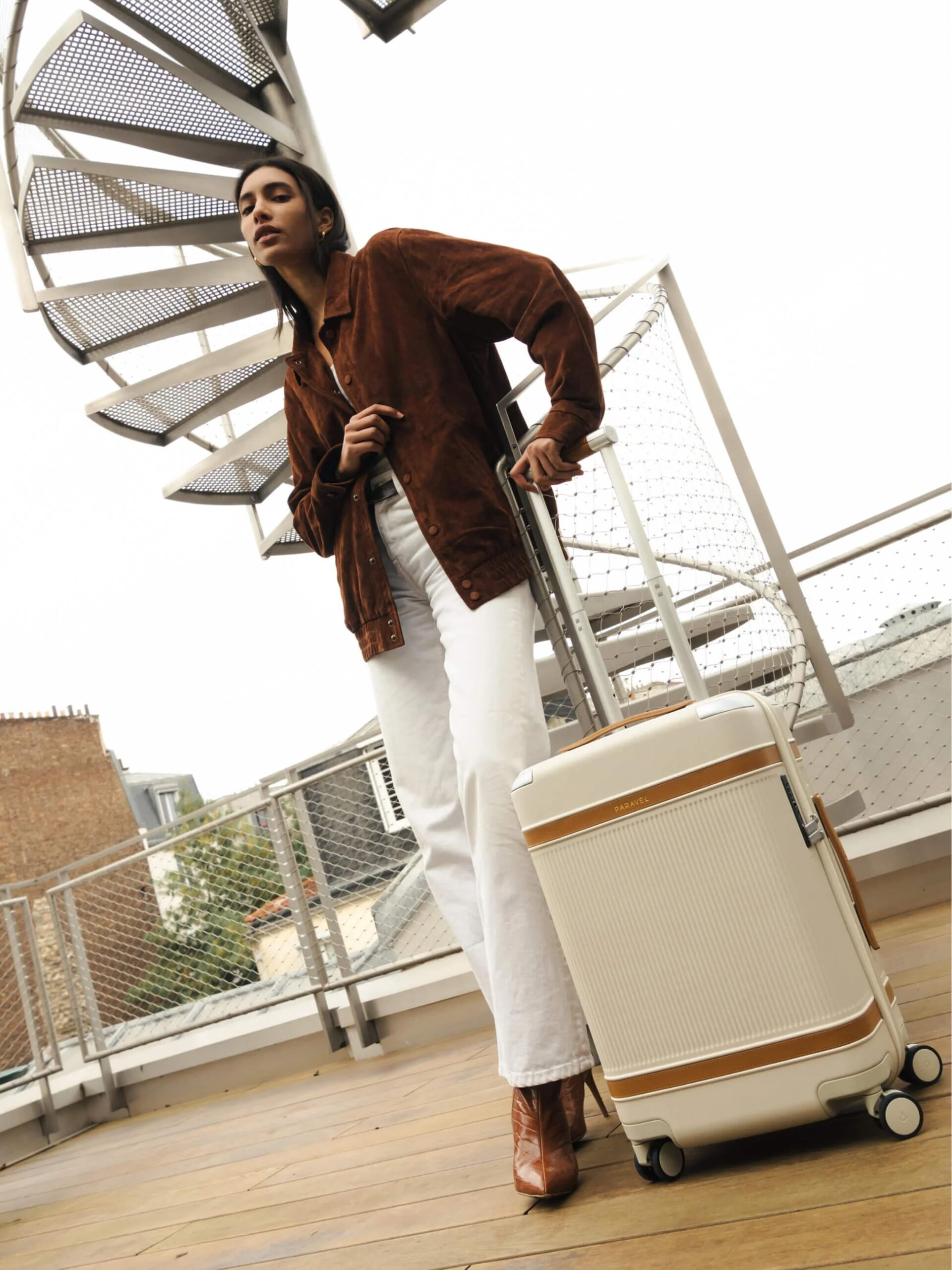 A person in a brown jacket and white pants stands on a wooden deck, leaning on a cream-colored suitcase, with a spiral staircase in the background.