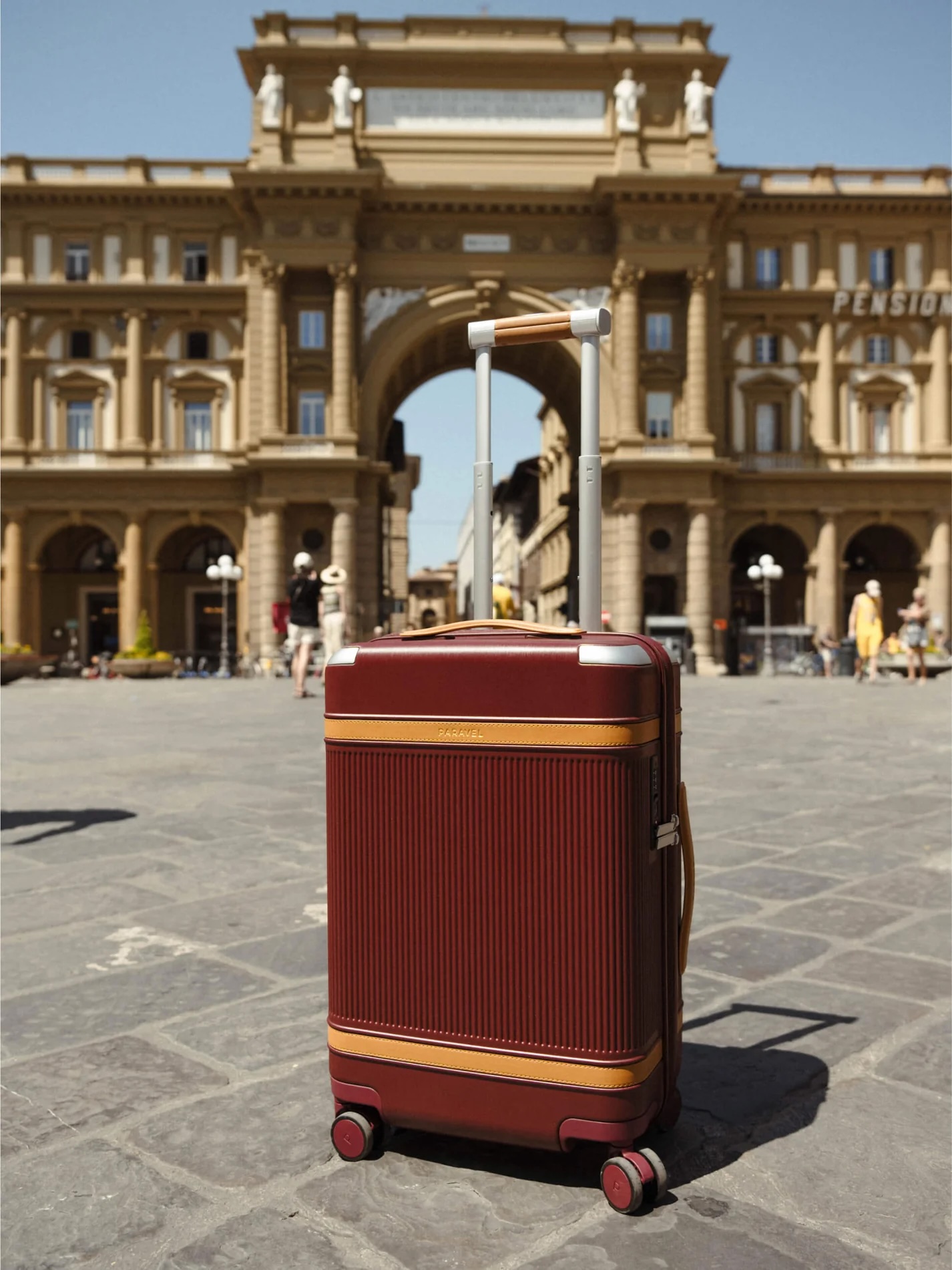 A red suitcase with extended handle is positioned on a cobblestone plaza in front of a historic archway and building under a clear blue sky.