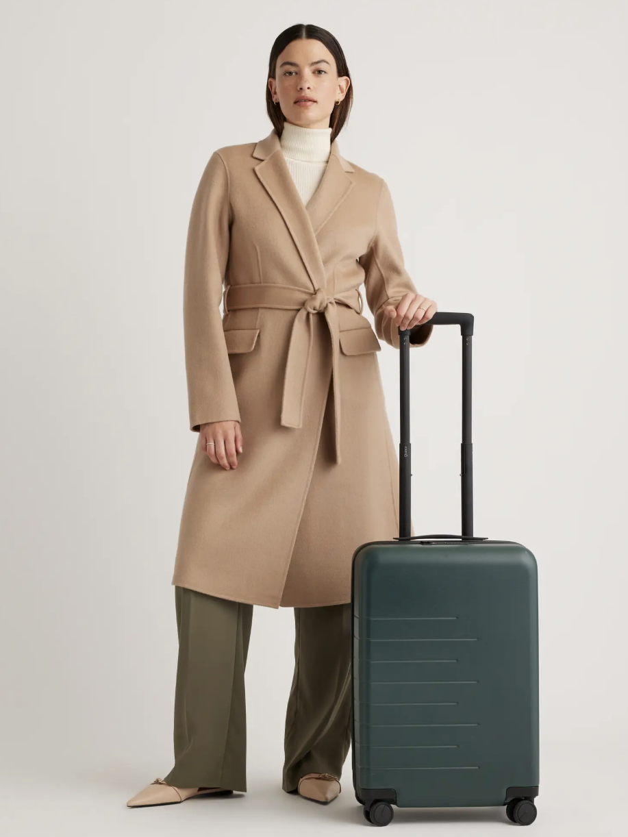 A person in a beige coat stands next to a dark green suitcase on wheels.