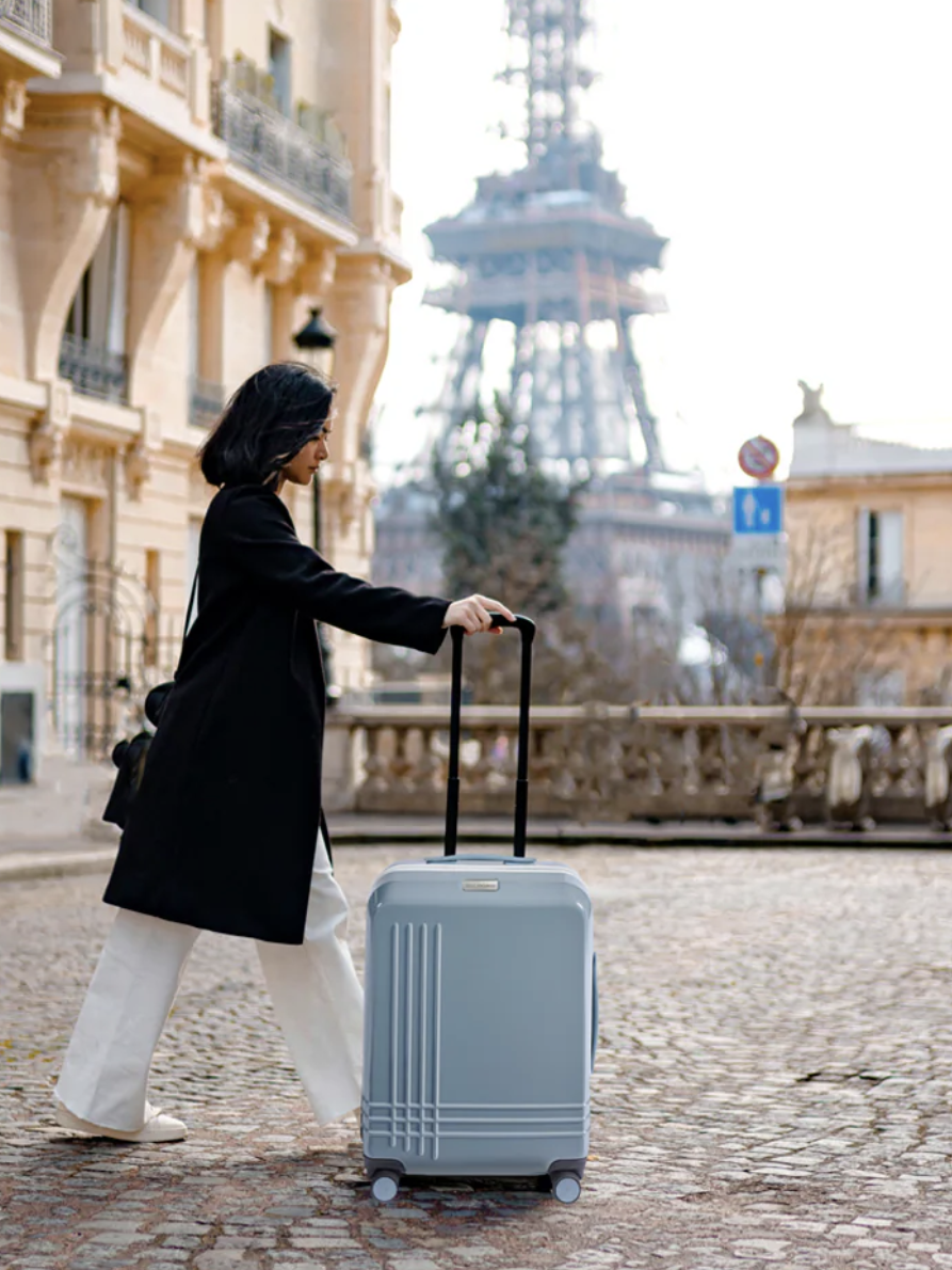 A person in a black coat and white pants walks on a cobblestone street with a gray suitcase. The Eiffel Tower and Parisian buildings are visible in the background.