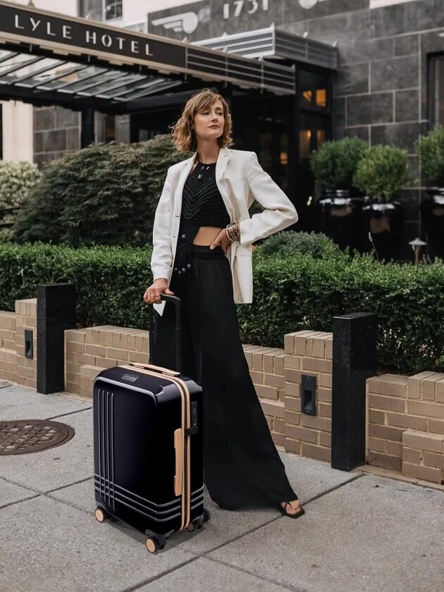 Person in a white blazer and black outfit stands on the sidewalk with a black suitcase in front of Lyle Hotel.