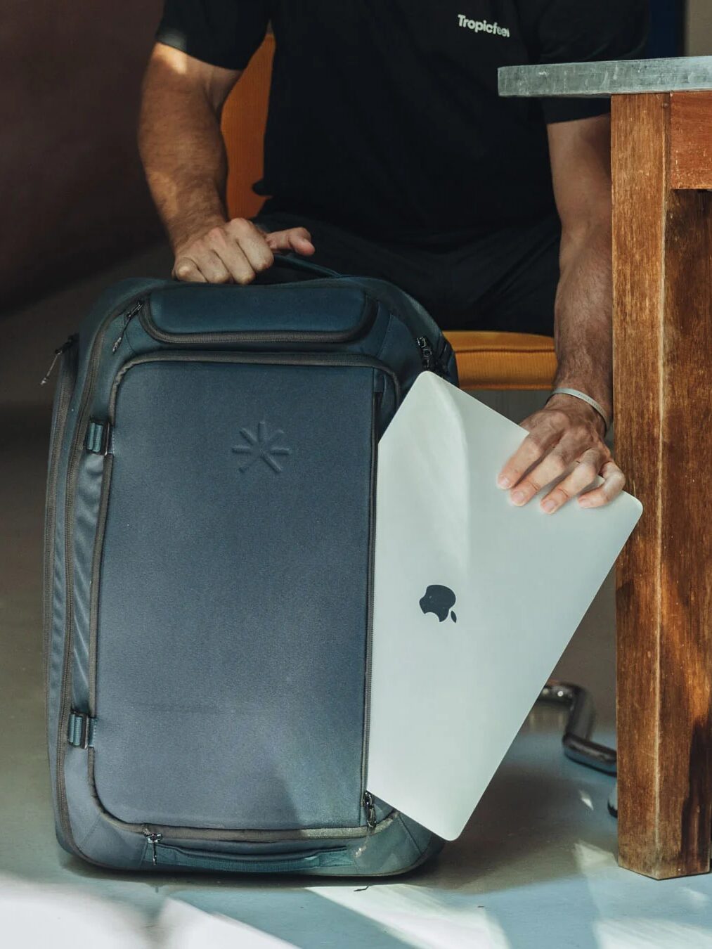 Person placing a silver laptop into a gray backpack beside a wooden table.