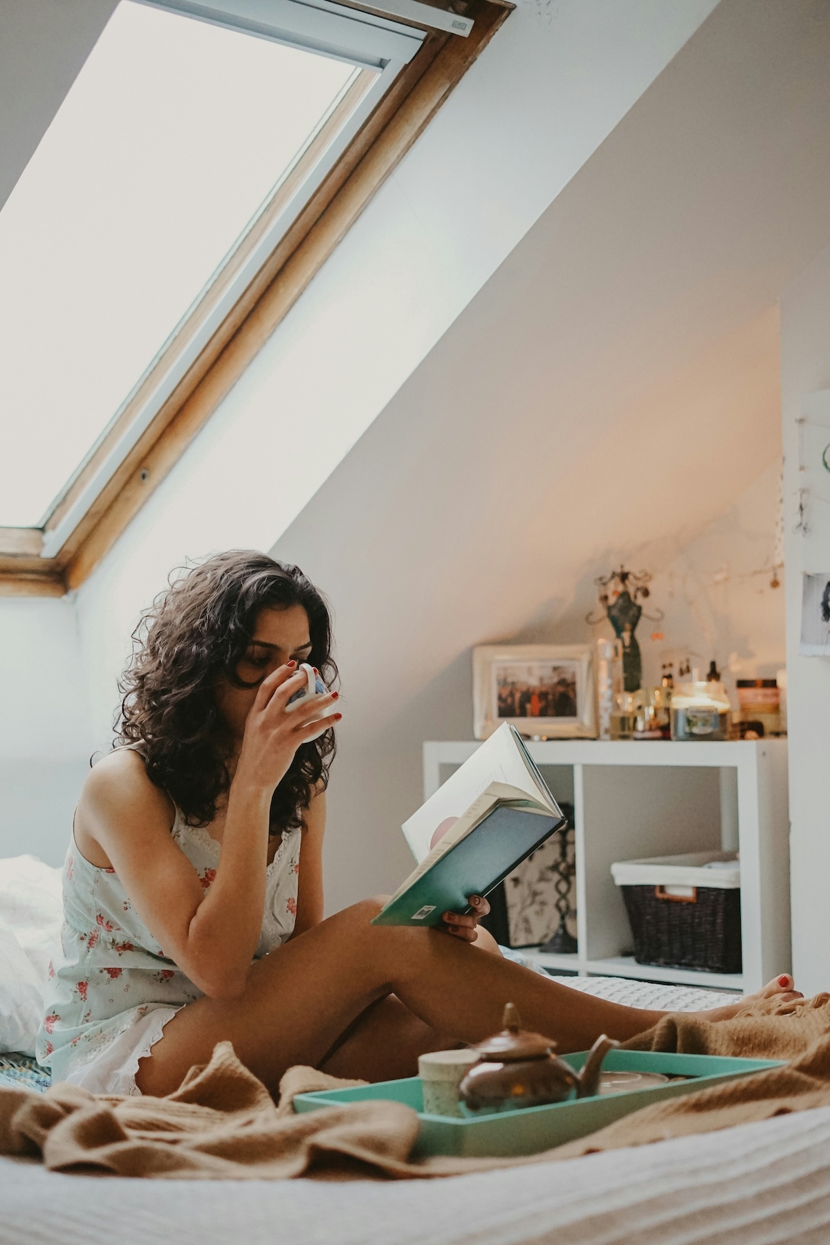 A person with curly hair sits on a bed near a skylight, drinking from a mug and reading a book. A teapot and various items are on the bed, with a shelf and picture frames in the background.