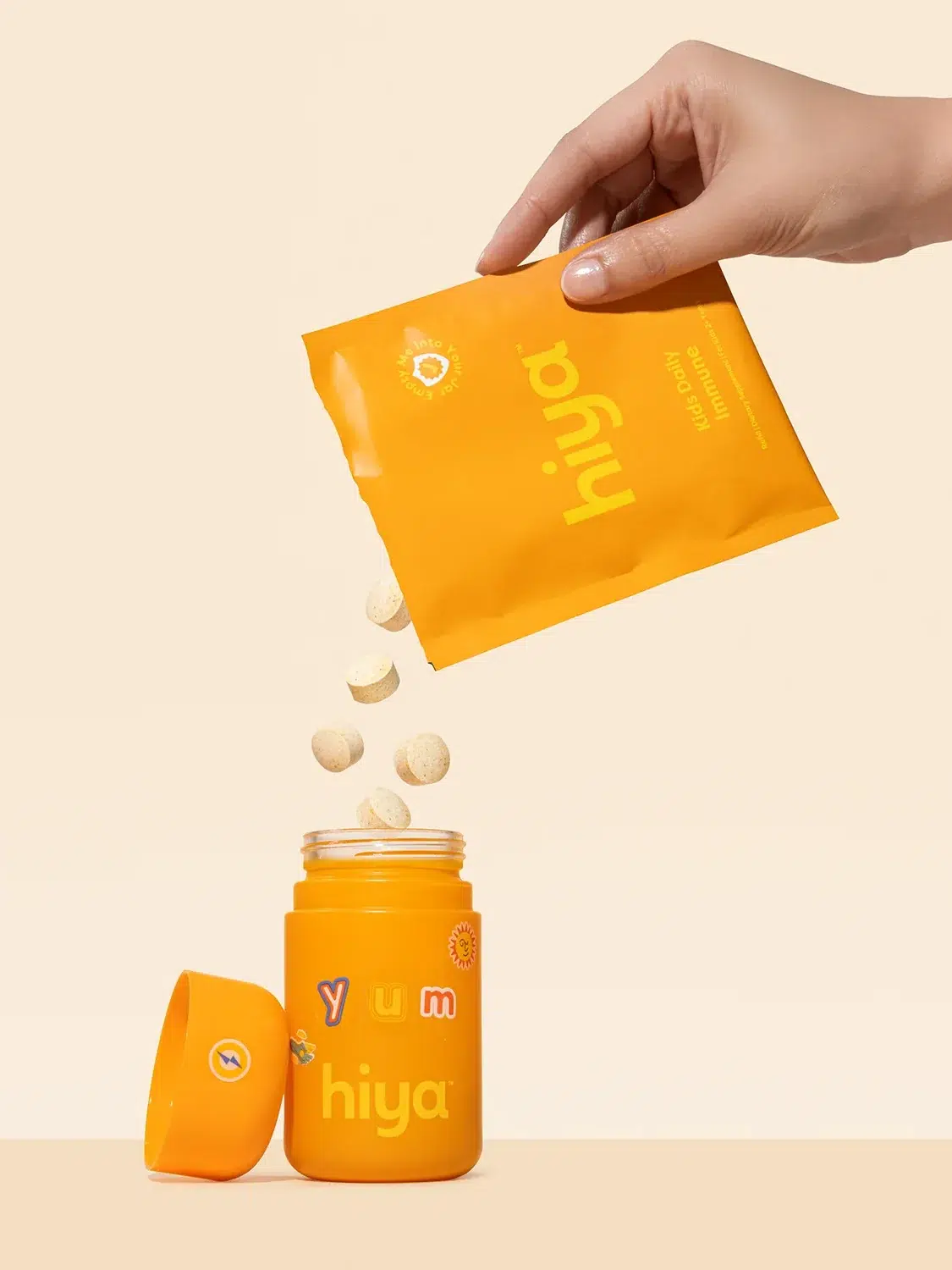 A hand pours tablets from an orange Hiya packet into an open orange jar labeled "Hiya" on a beige background. The jar's lid is to the left.