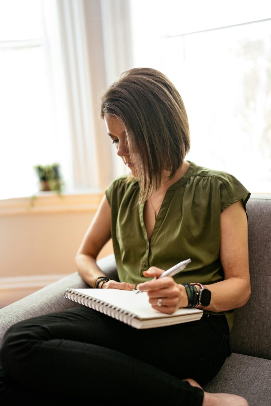 A person sits on a couch, writing in a notebook with a pen. They are wearing a green shirt and black jeans.