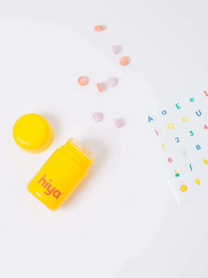 Yellow container labeled "hiya" with multicolored tablets scattered. Sticker sheet with letters, numbers, and symbols is nearby on a white background.