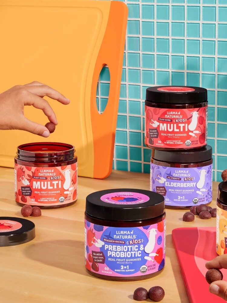 Four jars of Llama Naturals gummies on a kitchen counter with hands reaching for them. The jars are labeled Multi, Elderberry, and Prebiotic & Probiotic. A plant and orange cutting board are nearby.