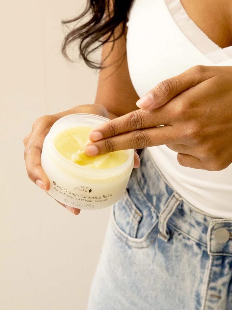 A person in a white top and jeans holds a jar of cleansing balm, using their fingers to scoop some out.