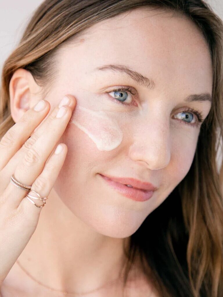 A woman applies a smear of skincare cream to her cheek, looking slightly off-camera. She has long hair and is wearing a ring on her left hand.