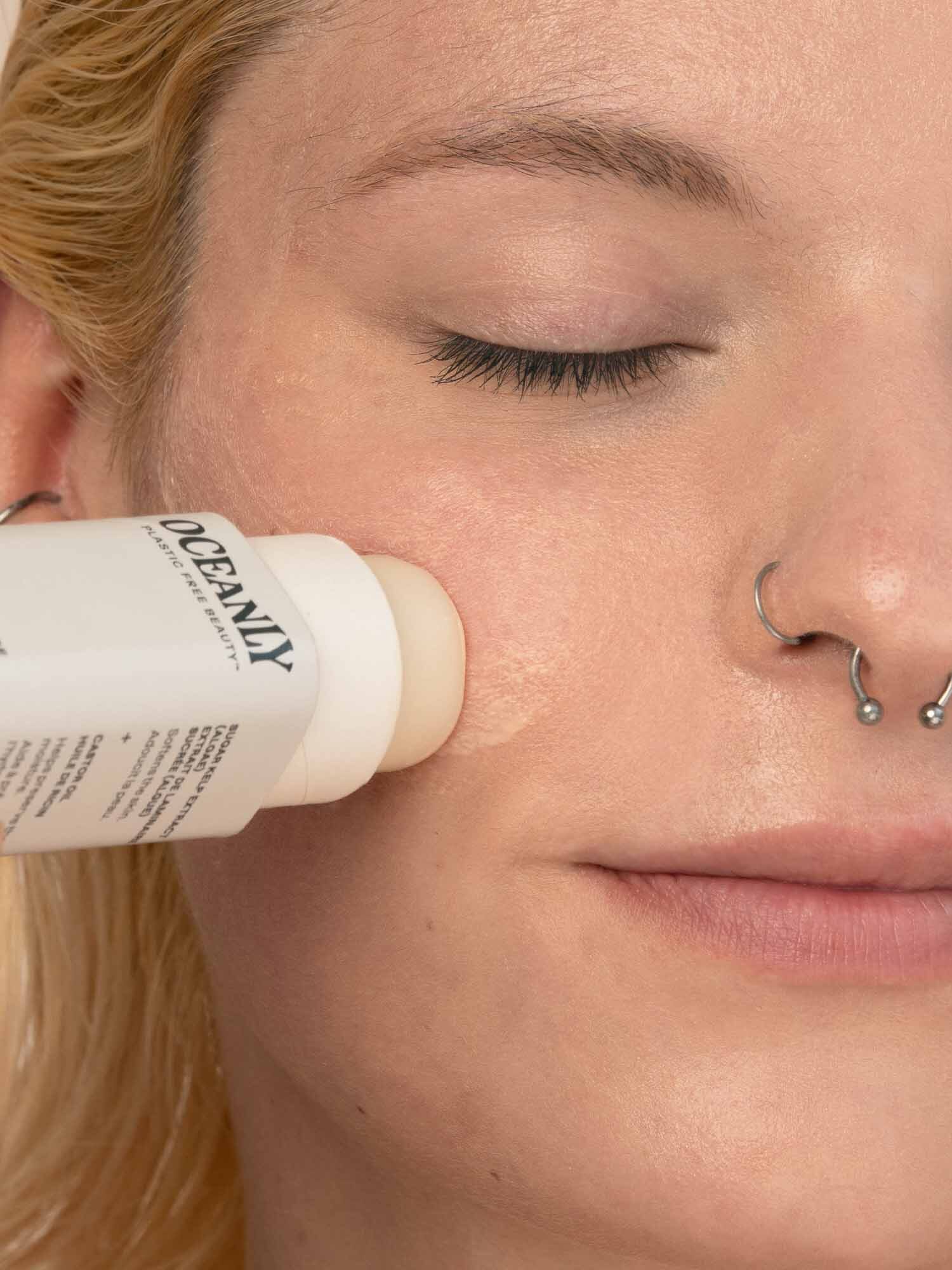 Person applying a stick product labeled "Oceamy" to their cheek, eyes closed. They have nose piercings and light blonde hair.