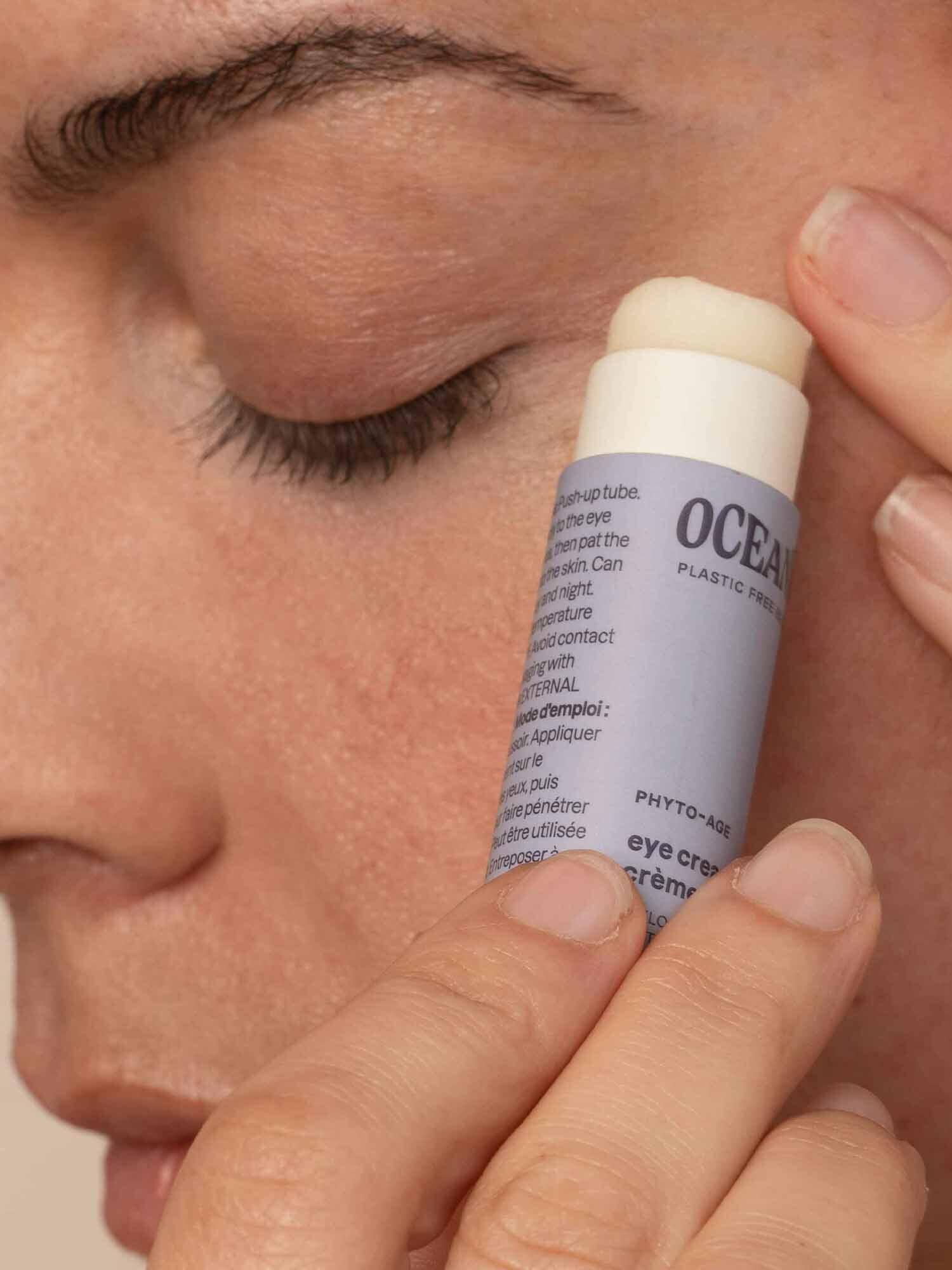 A person applies eye cream from a stick to their cheek, using their right hand. The cream stick has a light blue label.