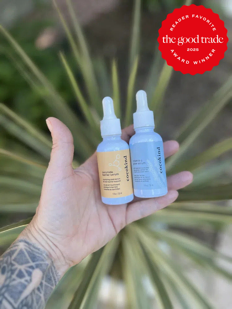 A tattooed hand holds two Cocokind serum bottles in front of a plant. A red badge reads "Reader Favorite: The Good Trade 2023 Award Winner.