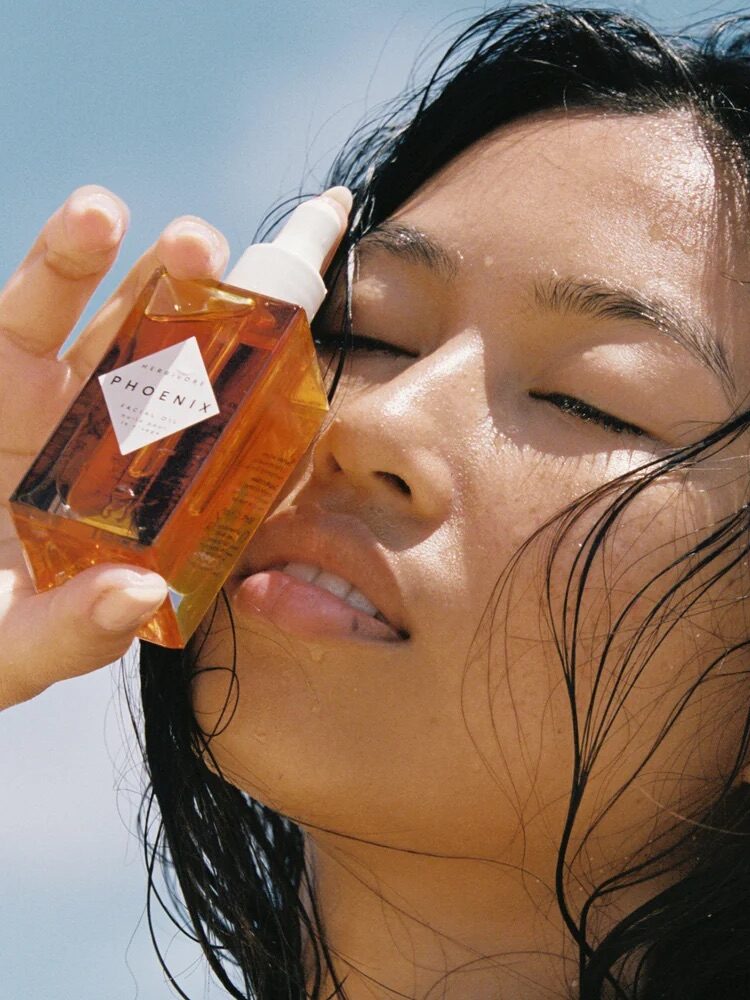 Person holding a dropper bottle labeled "Phoenix" near their face, with eyes closed, against a clear blue sky.