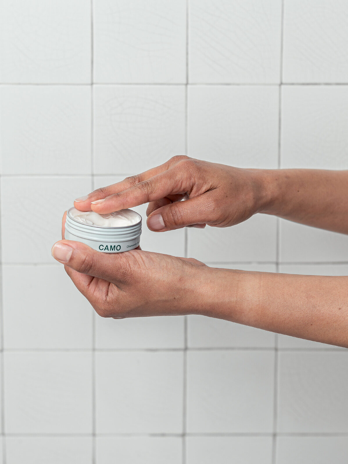 Hands holding a small, labeled jar of cream against a tiled background. A finger is about to touch the cream inside the open lid.