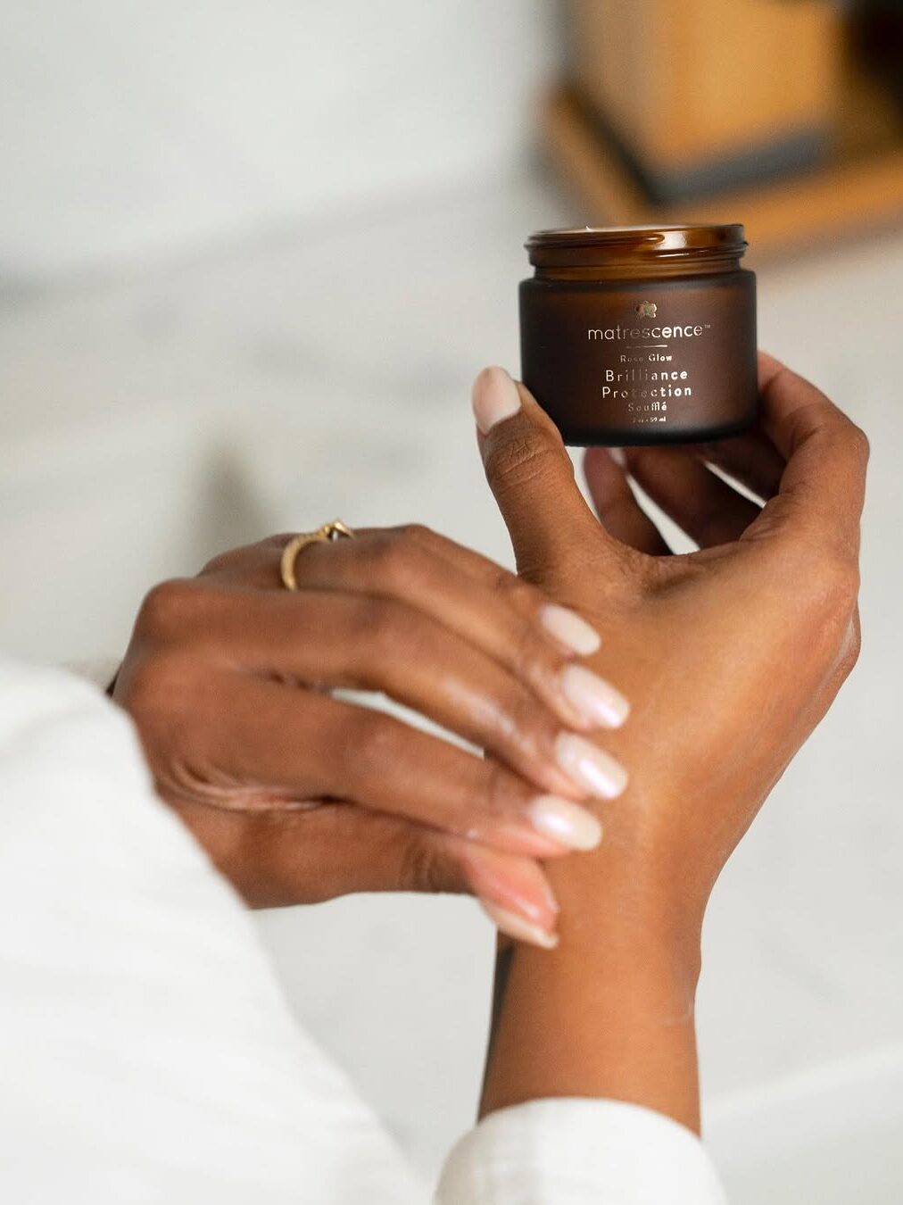 A person applies cream from a small brown jar labeled "maraesence brilliance protection" onto their wrist.