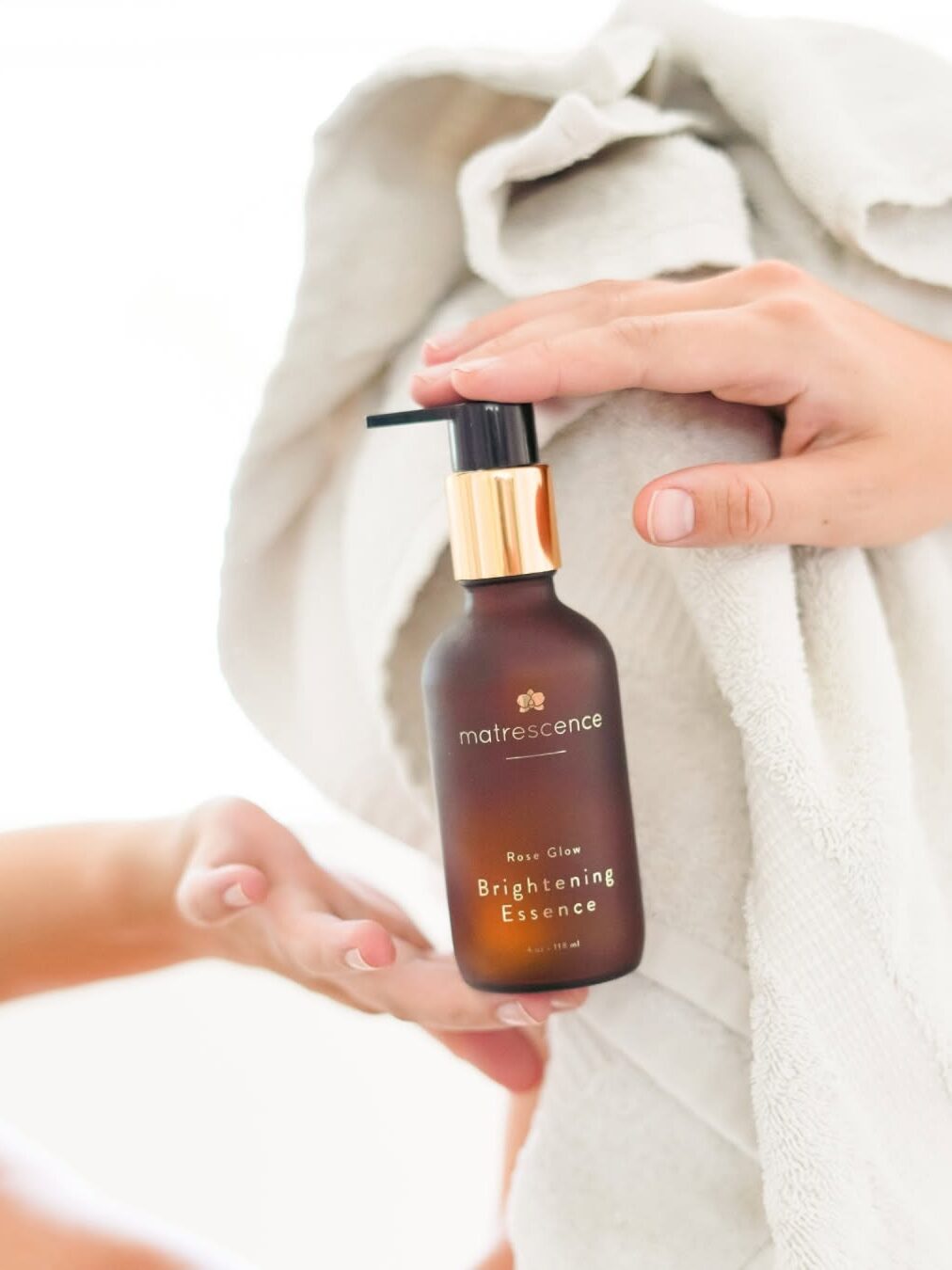 A person holds a dark brown bottle labeled "matrescence Brightening Essence" with a hand pump, next to a white towel.