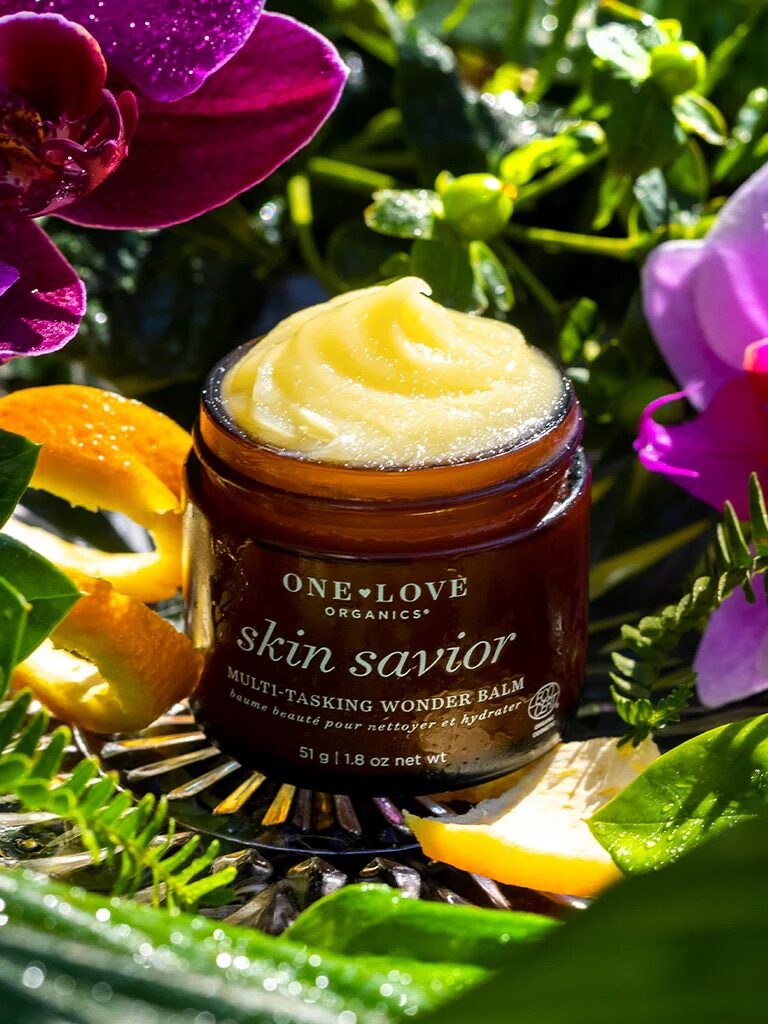 A jar of One Love Organics Skin Savior balm surrounded by vibrant pink orchids, ferns, and orange peels. Sunlight highlights the product against the lush greenery backdrop.