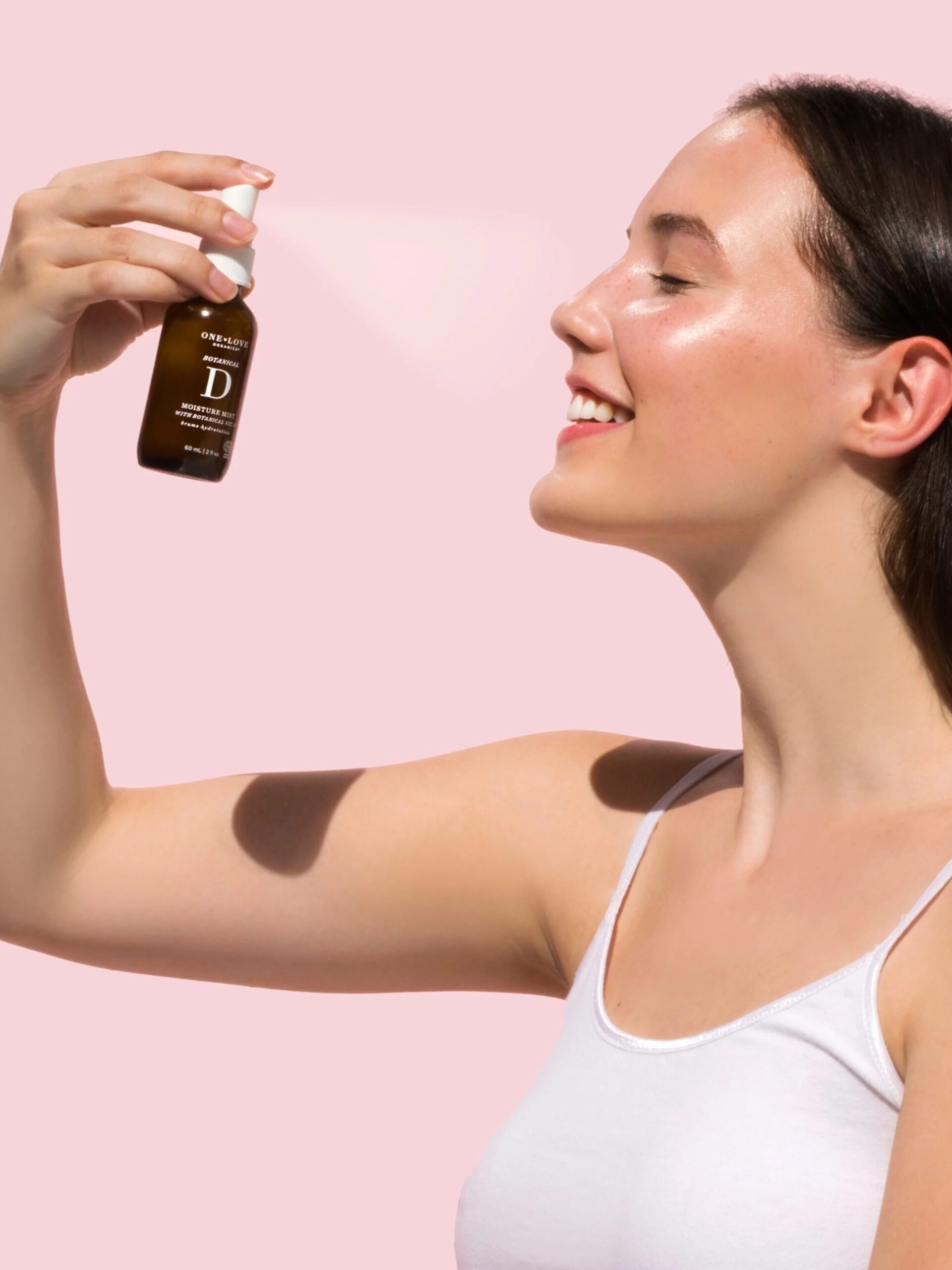 A woman with long dark hair sprays a serum labeled "Vitamin D" onto her face against a pink background. She is wearing a white tank top and smiling.