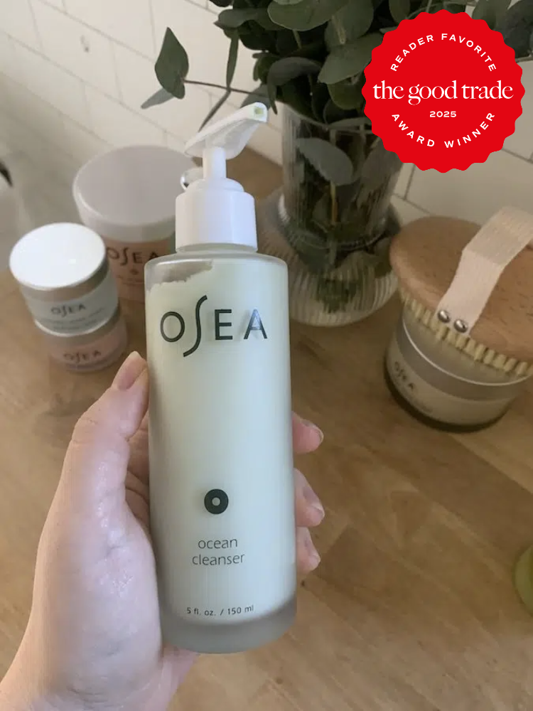 A hand holding a bottle of OSEA Ocean Cleanser is in focus. In the background, there are other skincare items and a plant. A red badge on the top right corner reads: "The Good Trade 2023 Award Winner.