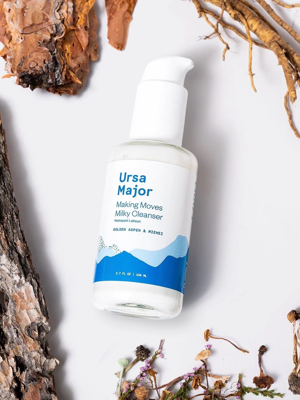 A bottle of Ursa Major Making Moves Milky Cleanser surrounded by dried plants and bark on a white background.