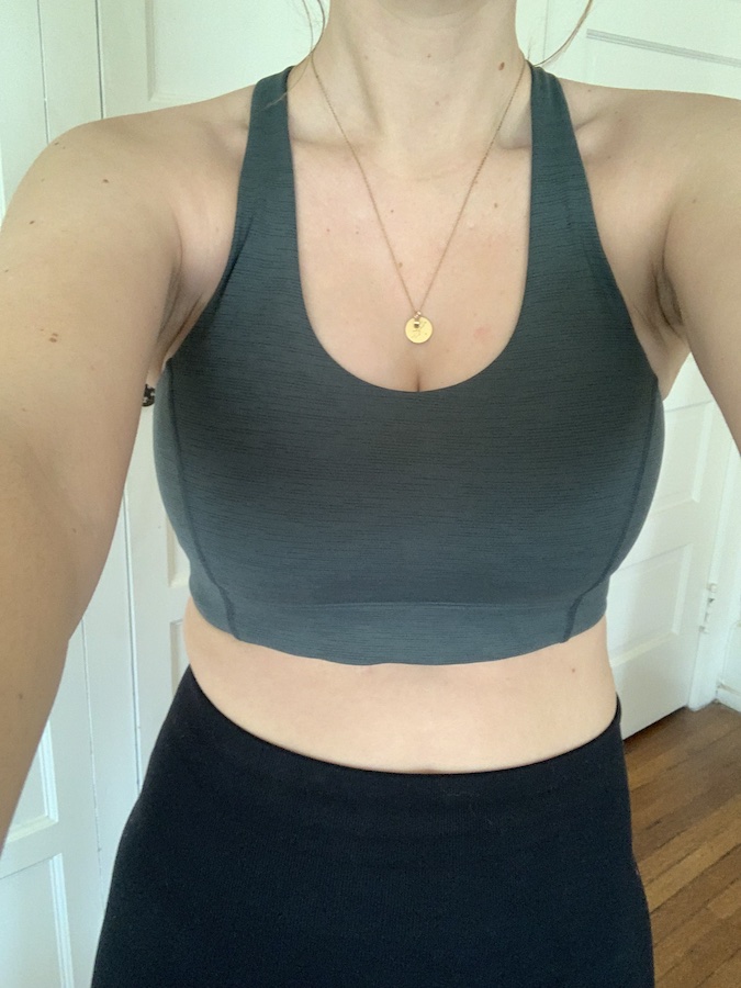 Person wearing a gray sports bra and black leggings, with a gold necklace, standing indoors against a white background.