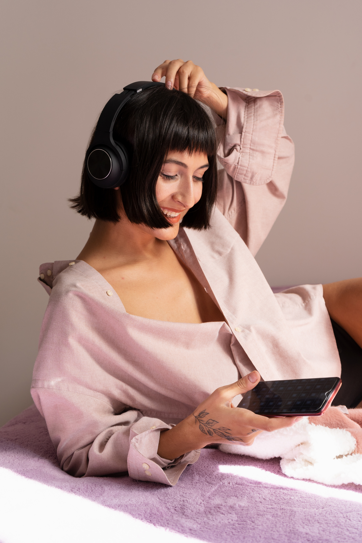 Woman with headphones smiles while looking at a smartphone, dressed in a light pink shirt, sitting on a purple surface with a neutral background.