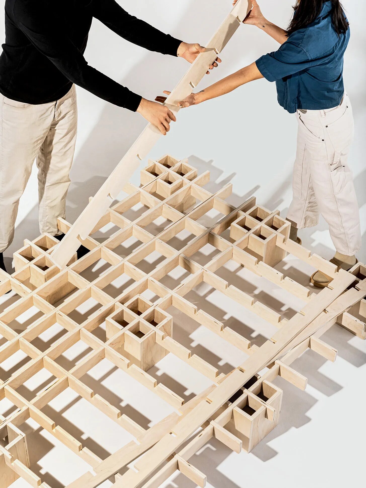 Two people are assembling a wooden lattice-like structure on a white background.