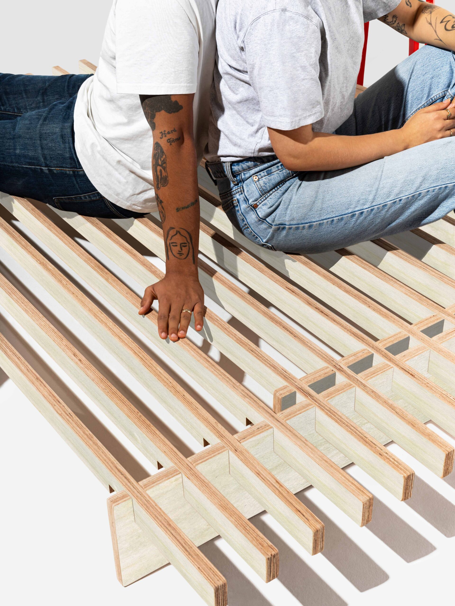 Two people sit on a modern wooden bench with open slats. Both wear casual clothing and display visible arm tattoos.