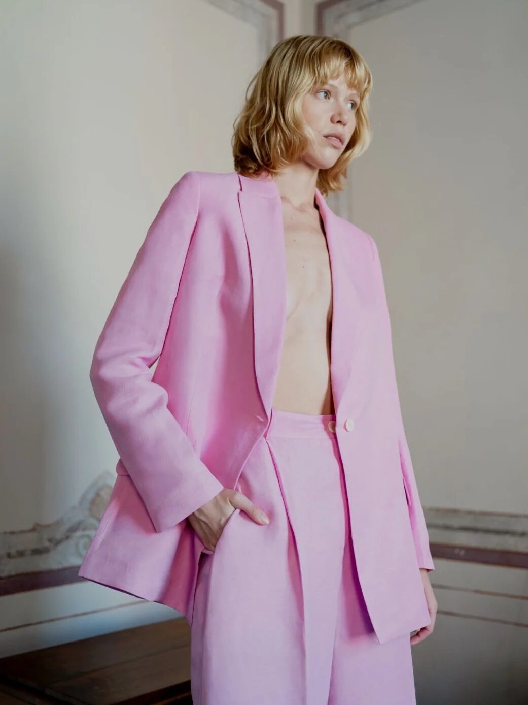 Person wearing a light pink suit with no shirt underneath, standing in a room with soft lighting.