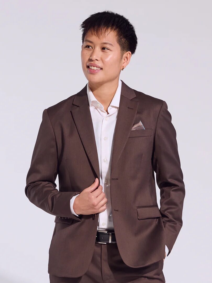 A person in a brown suit and white shirt stands against a plain background, adjusting their jacket with one hand in the pocket.