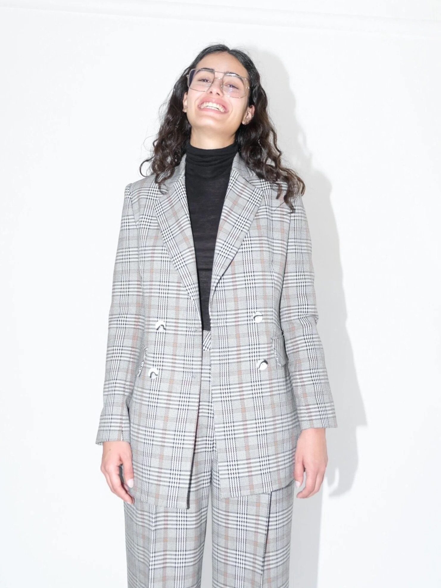 A person wearing a checked suit and glasses stands against a white background, smiling.