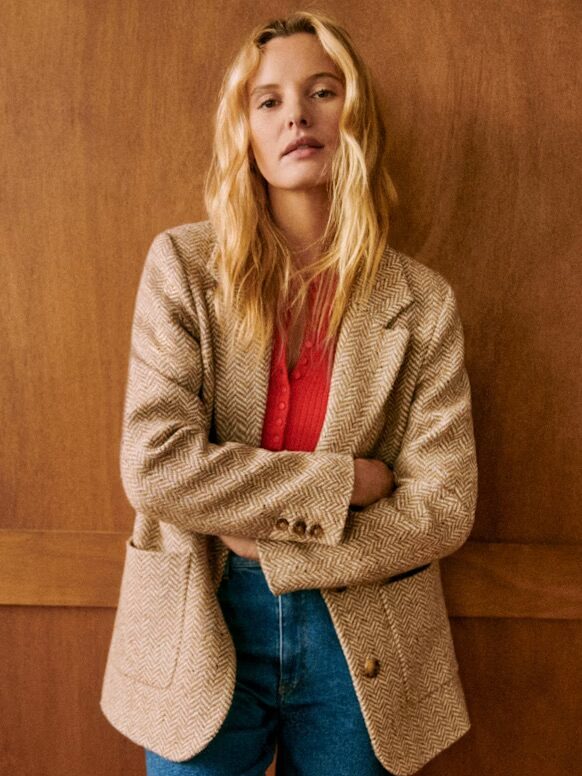 Person with long hair wearing a beige herringbone blazer, red top, and blue jeans, standing with arms crossed in front of a wooden wall.