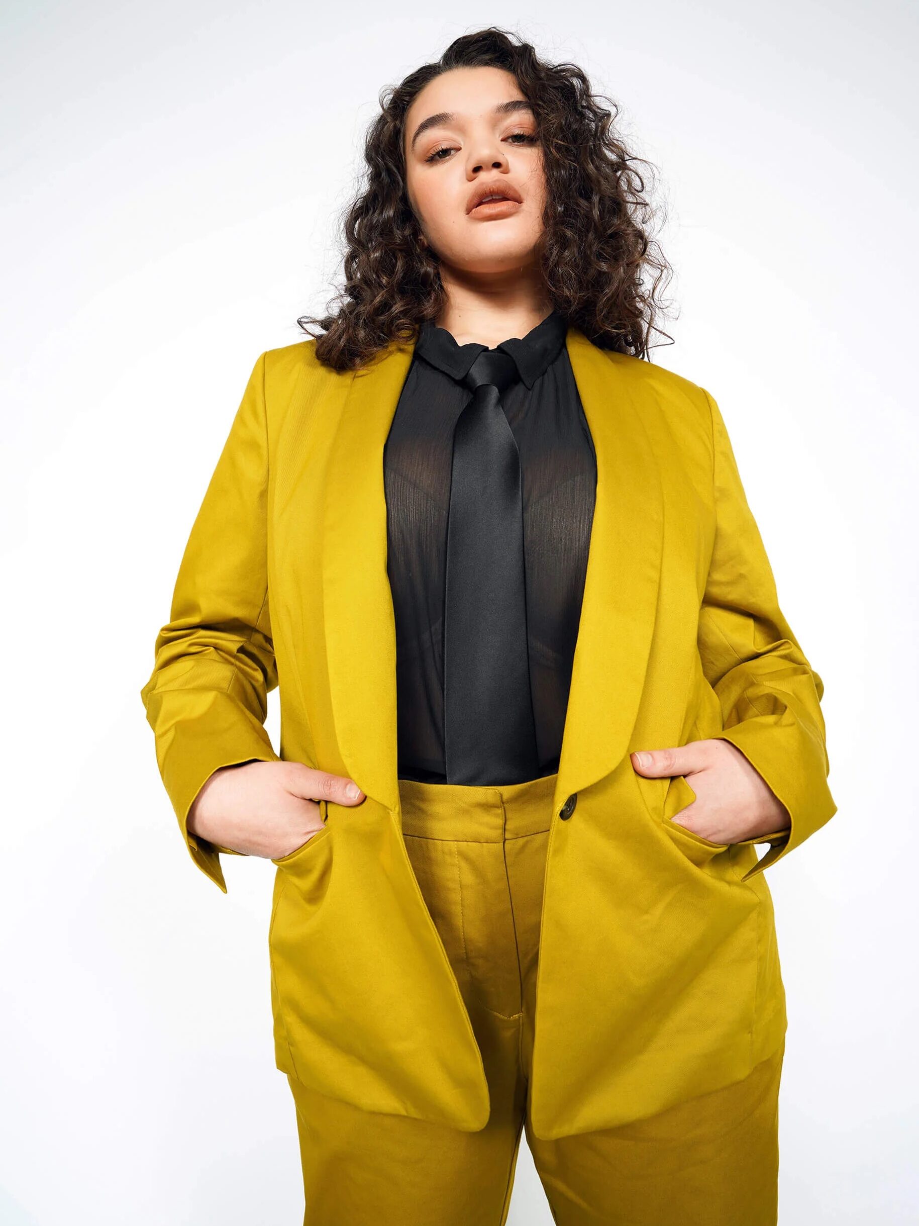 Person posing confidently in a mustard yellow suit with hands in pockets, wearing a black shirt and tie.