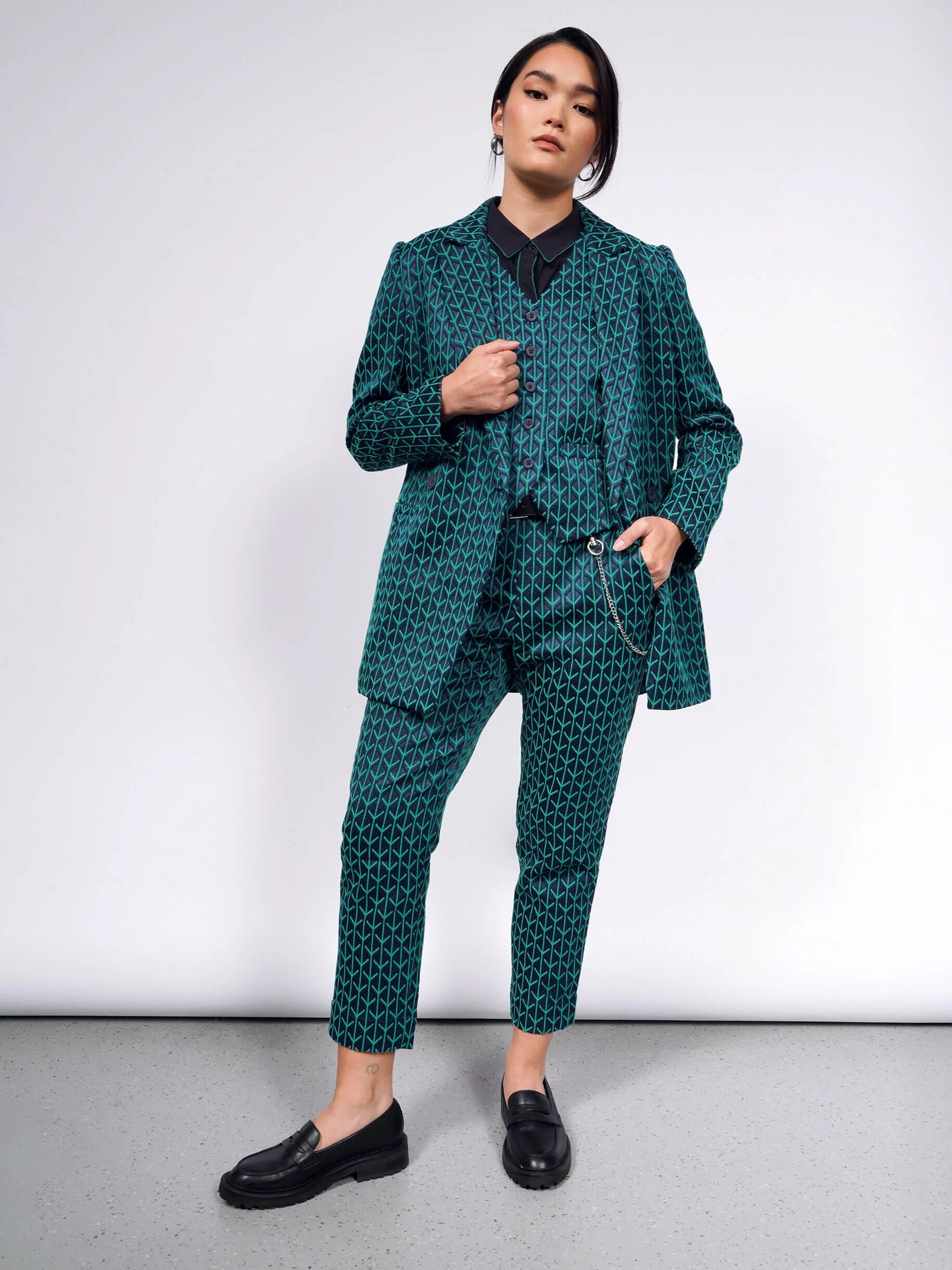 Person wearing a patterned teal suit with a black shirt and black loafers, standing against a plain background.