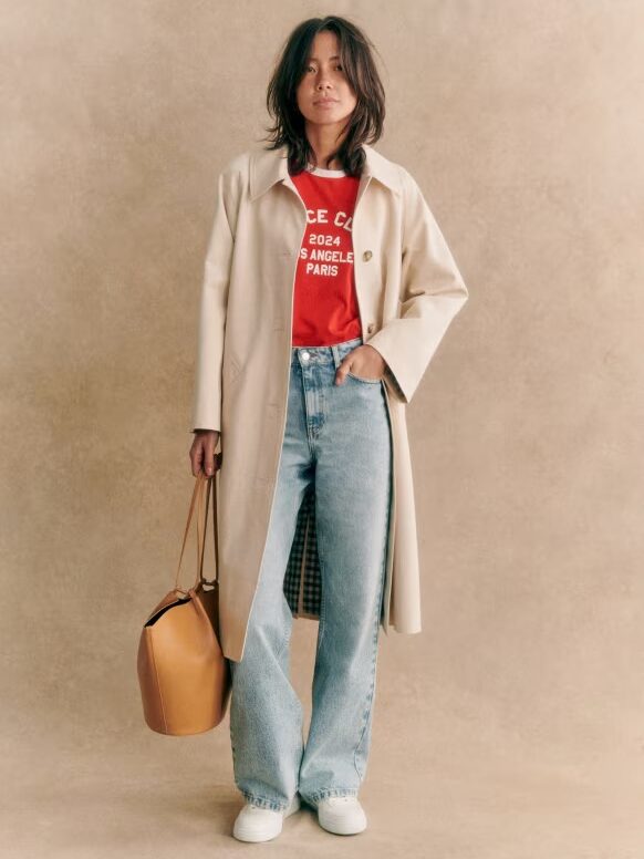 Person wearing a long beige coat over a red shirt with text, paired with blue jeans and white shoes, holds a brown handbag against a plain backdrop.