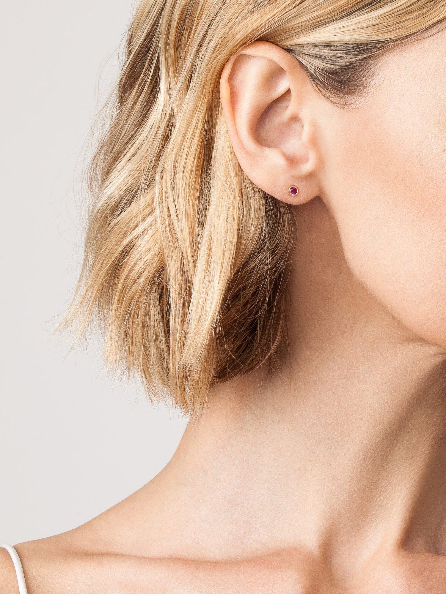 A woman with short blonde hair wears a minimalist stud earring, showing her ear and part of her neck against a plain background.