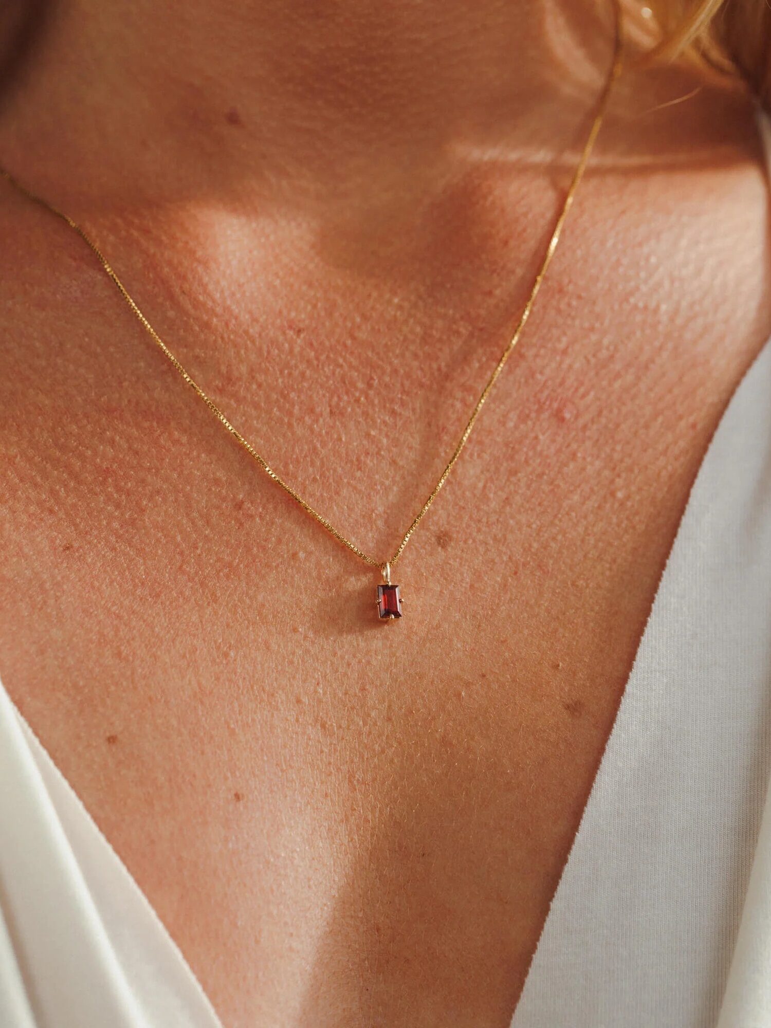 A gold necklace with a rectangular red pendant is worn by a person in a white V-neck top.