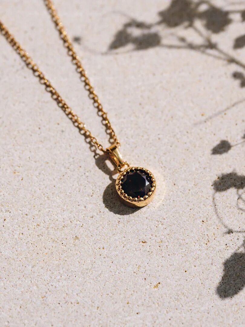 A gold necklace with a round black pendant on a textured beige surface, with plant shadows nearby.