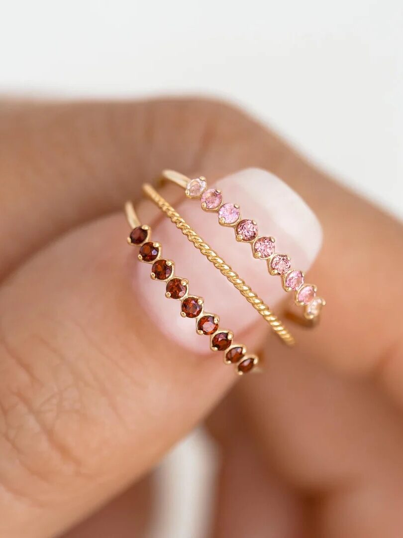 Three delicate rings with gemstones in red and pink hues held between fingers against a blurred background.