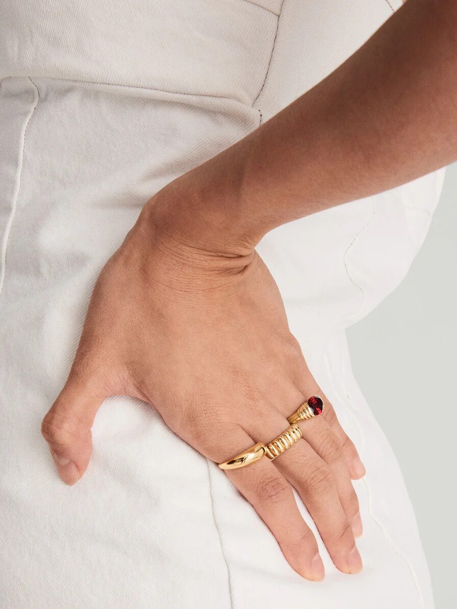 Close-up of a hand resting on a hip, wearing two gold rings, one featuring a red gemstone. The person is dressed in a white outfit.