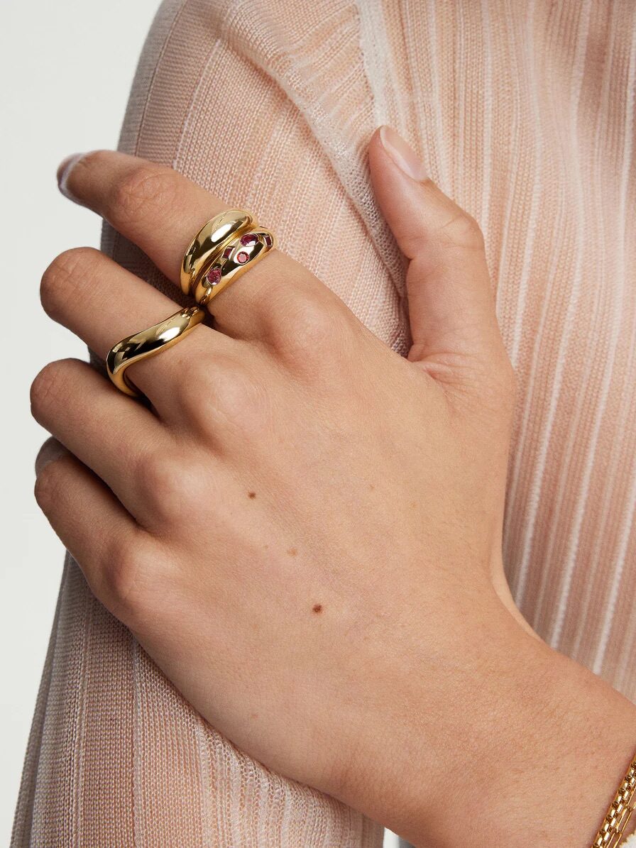 A hand wearing two gold rings on the index and middle fingers, with a pink gem on one ring, and a gold bracelet on the wrist. The background is a light-colored, ribbed fabric.