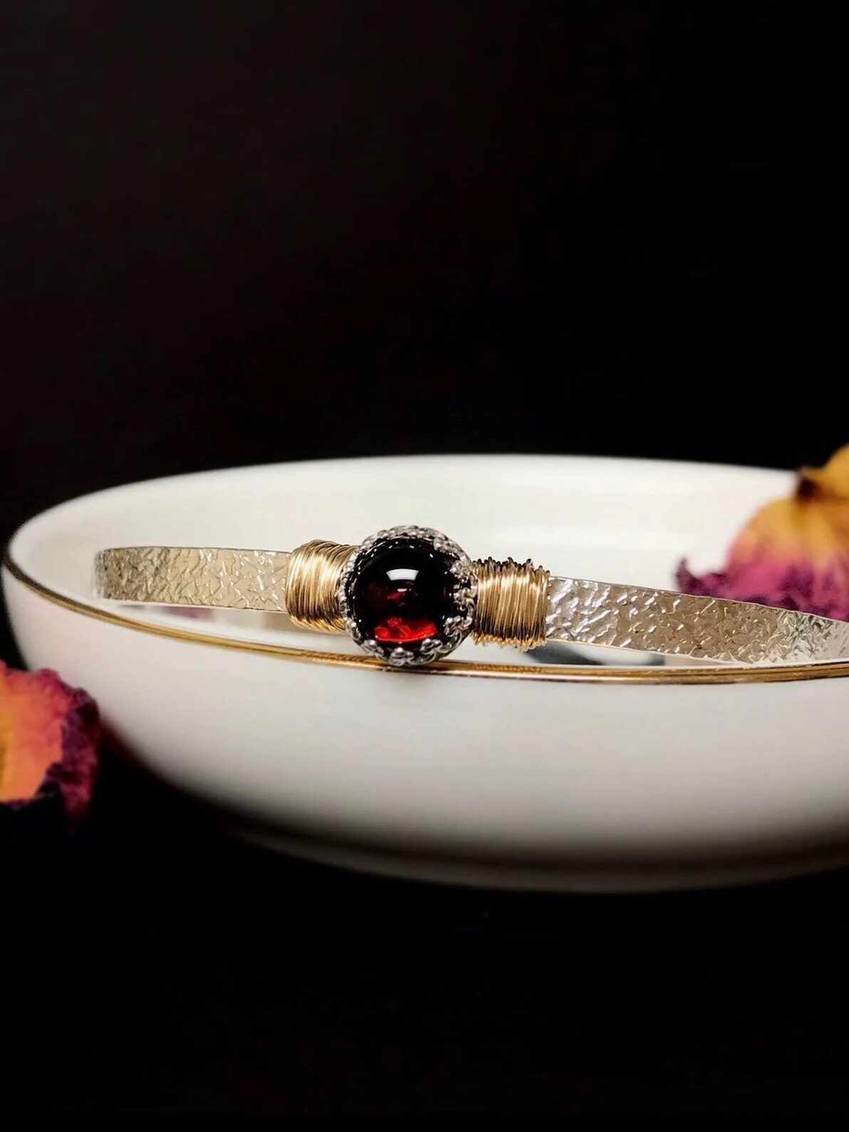 A silver bracelet with a central red gemstone rests on a white bowl, surrounded by dried pink and yellow petals.