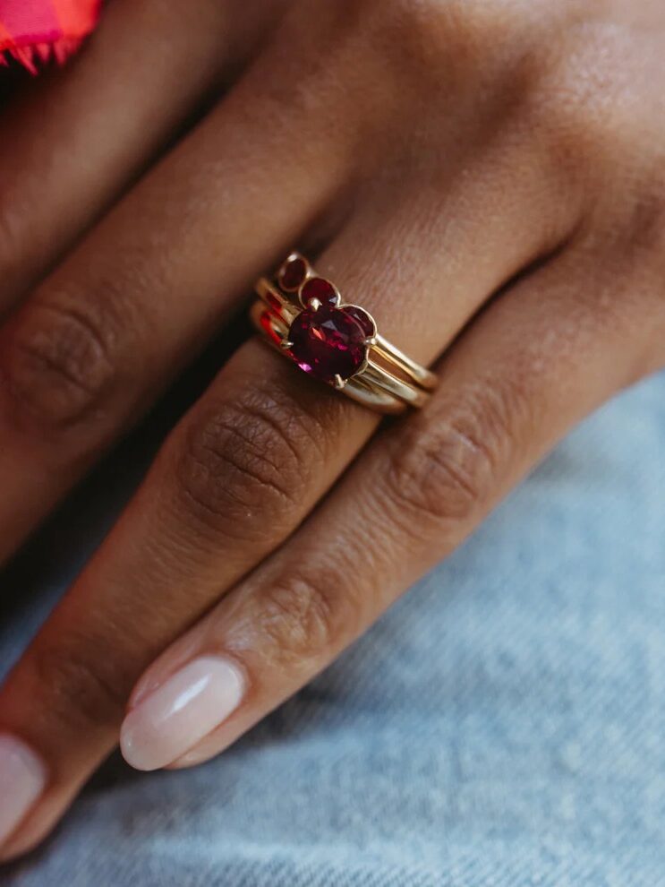 A hand wearing a gold ring with a large red gemstone and two smaller stones above it, resting on denim fabric.