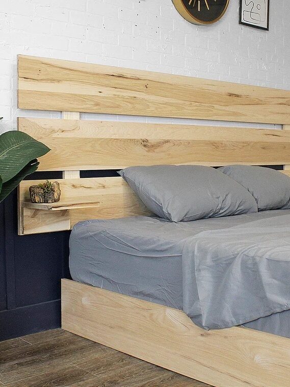 A bed with gray bedding, a wooden headboard with shelves, and potted plants on each side in a room with a white brick wall and a wall clock.